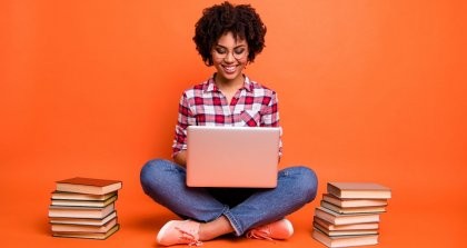 Eine junge Frau ist von Büchern umgeben und sitzt schreibend an ihrem Laptop.